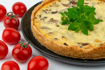 Closeup wide studio shot of freshly baked yellow French salty cake, or quiche, with mushrooms, red cherry tomatoes, on a black design plate, isolated on white background.