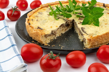 Closeup wide studio shot of freshly baked yellow French salty cake, or quiche with mushrooms, red cherry tomatoes, green mint and kitchen cloth, on a black design plate, on white background