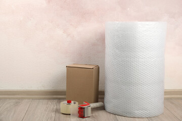 Bubble wrap roll, tape dispenser and cardboard box on floor near pink wall. Space for text