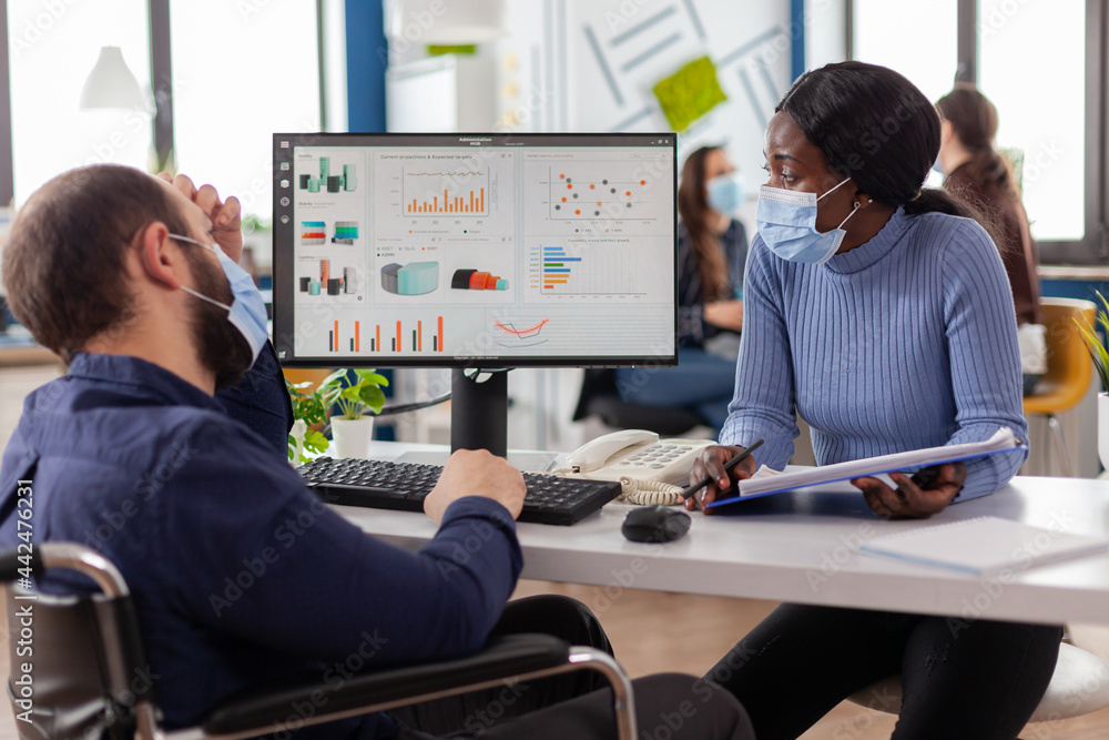 Poster paralysed freelancer with face mask discussing with black woman partner financial problems, checking