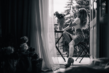 A young girl in the morning in the doorway of the balcony sits at a table with flowers