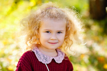 A large portrait of a small blonde girl in a burgundy sweater in a sunny park in autumn. The child smiles and looks at the camera.