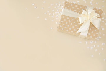 Beautiful pastel colored beige Christmas gift wrapped in kraft dotted paper with white ribbon and bow on beige background and circle confetti. Neutral feminine minimal present background, copy space