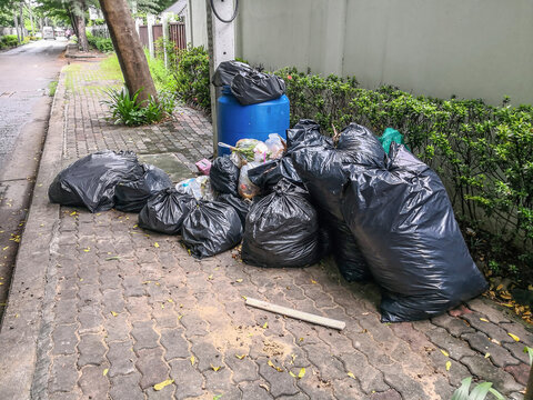 Black garbage bag pile near municipal trash bin full on sidewalk.
