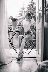 A young girl in the morning in the doorway of the balcony sits at a table with flowers