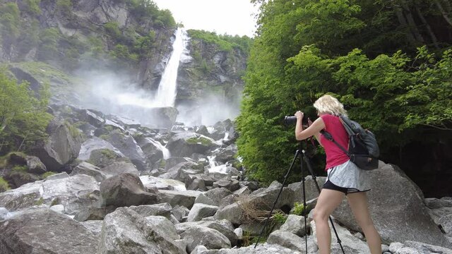 Woman photographer taking pictures with tripod at the Foroglio waterfall over the Bavona River in Foroglio town of Switzerland. Bavona river leading to Lake Langensee.