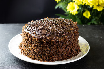Cake Prague on a black table against the yellow chrysanthemums. View from the side. Chocolate cake on a white plate