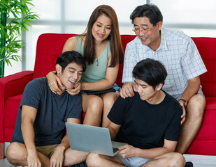 Fototapeta premium Four members Asian family consisting of parents, two adult sons happily spending time together during holidays, using laptop in comfortable living room at home. Presenting modern technology concept.