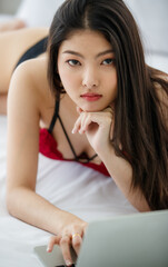 Naklejka premium Beautiful young naughty sexy girl with long hair and romantic underwear lie down on bed and attentively consider to type on laptop on rest day, new normal work at home concept.