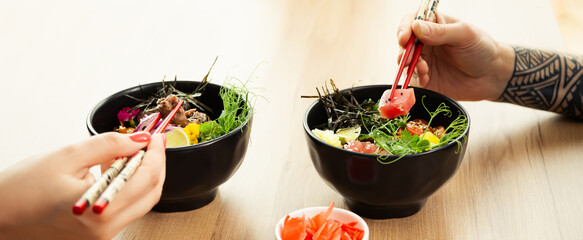 Man and woman eating poke salad with chopsticks. Dab tuna salad in a bowl. People in the restaurant...