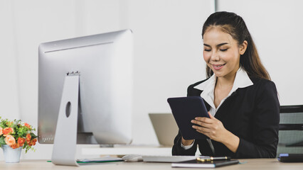Asian office people are businesswoman wearing black suit using tablet instead computer at work desk look to feel happy with smile on face