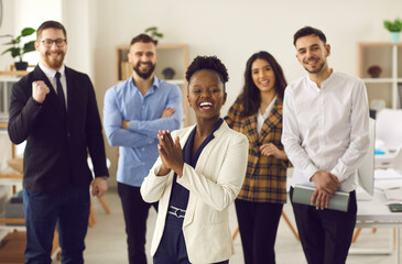 Multiethnic team celebrating success. Happy business leader or manager applauding standing in front of employees in modern office. Black woman clapping hands with multiracial colleagues in background