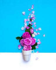 Creative layout with pouring fresh rose flowers and leaves into tea cup on pastel blue background. Creative herbal tea concept. Flat lay.