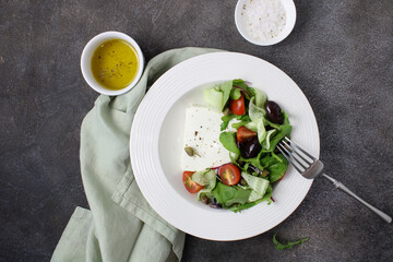 Vegetable salad recipe with cheese, tomatoes, cucumbers, red onions, arugula and olives in a white plate on a dark background, top view