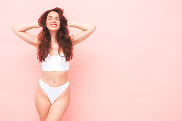Portrait of young beautiful smiling woman in white lingerie. Sexy carefree cheerful model in underwear posing near pink wall in studio. Positive and happy female enjoying morning.Closed eyes