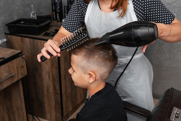 Hairdresser dries hair with a hairdryer to a little boy. The process of creating a fashionable hairstyle.