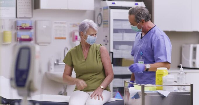A Wide Shot Of An Australian Male Medical NurseAdministering A Covid-19, SARS-CoV-2 Vaccine Injection With A Needle and Syringe, To An Elderly Woman. Wearing Safety Gloves And Protective Mask