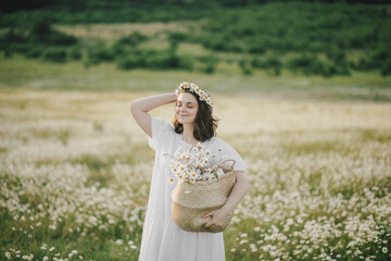 Fototapeta na wymiar Young woman wearing white dress holding straw basket with flowers on chamomile field.