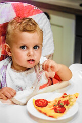 little baby boy sits on a chair and a slice of pizza on a plate. unhealthy food