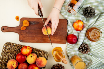 top view of female hands cutting fresh sweet peaches on a wooden cutting board and a glass of honey with dried apricots on white background