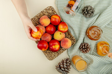 top view of fresh ripe peaches and nectarines on a wicker tray and glass jars with peach jam honey and dried apricots on plaid fabric on white background