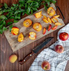 top view of halves of fresh ripe peaches on a wooden cutting board with kitchen knife and green leaves on rustic background