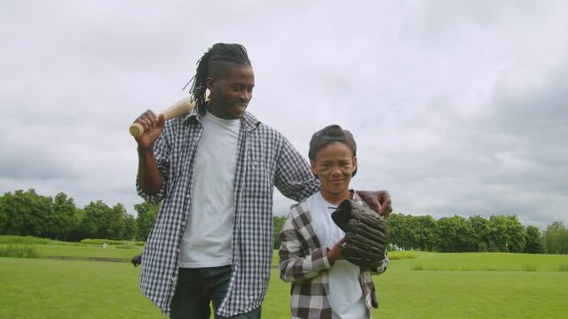 Cheerful handsome african american father with baseball bat on shoulder hugging cute happy preadolescent son with ball and baseball glove, walking along green field and chatting after baseball game.
