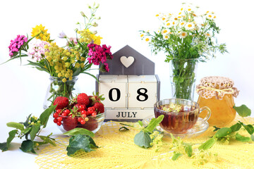Calendar for July 8 : the name of the month of July in English, cubes with the numbers 0 and 8, bouquets of wild flowers, jam, fruit, a cup of tea on a yellow openwork napkin