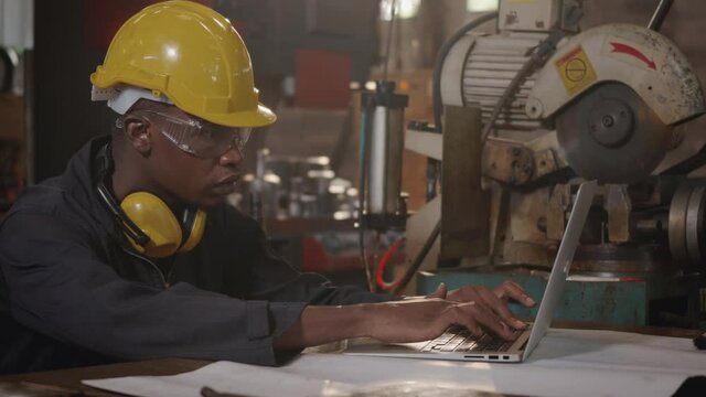 American industrial black young worker man with yellow helmet and ear protection typing keyboard of laptop computer in front machine, Confident engineer at work in the industry factory
