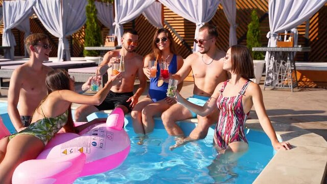 Multiracial group of friends toasting, clinking glasses with cocktails at swimming pool party. Happy young people in swimwear dancing, clubbing with inflatable flamingo, mattresses in luxury resort.