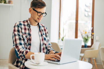 young man wearing glasses, uses laptop for video call on laptop computer, sits in a cafe and drinks a cup of coffee