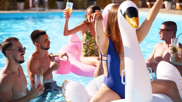 Friends having party with cocktails in holiday villa swimming pool. Happy young people in swimwear dancing, clubbing with inflatable flamingo, swan, mattress in luxury resort on sunny day. Slow motion