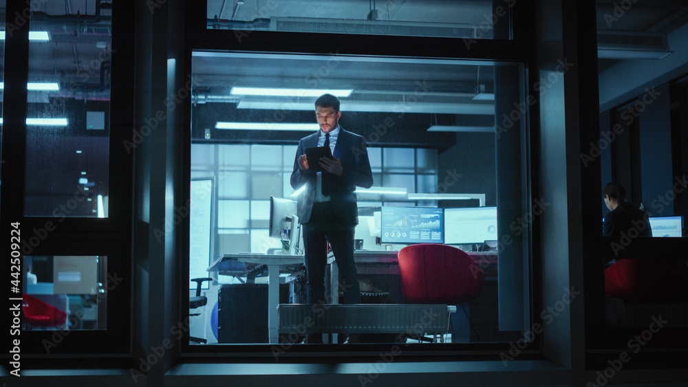 Poster Shot from Outside: Businessman Working on a Digital Tablet Computer in the Office. Manager Checking Emails while Standing by the Window. Employee Planning Financial Tasks and Business Development.