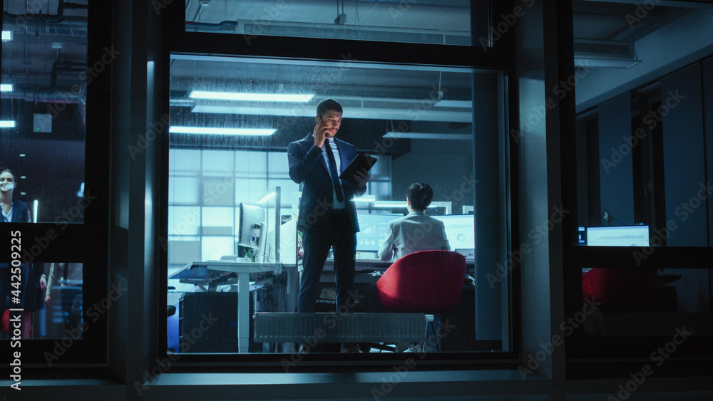 Poster Shot from Outside: Businessman Working on a Tablet Computer in the Office. Manager Talks on the Mobile while Standing by the Window. Employee Planning Financial Tasks and Business Development.