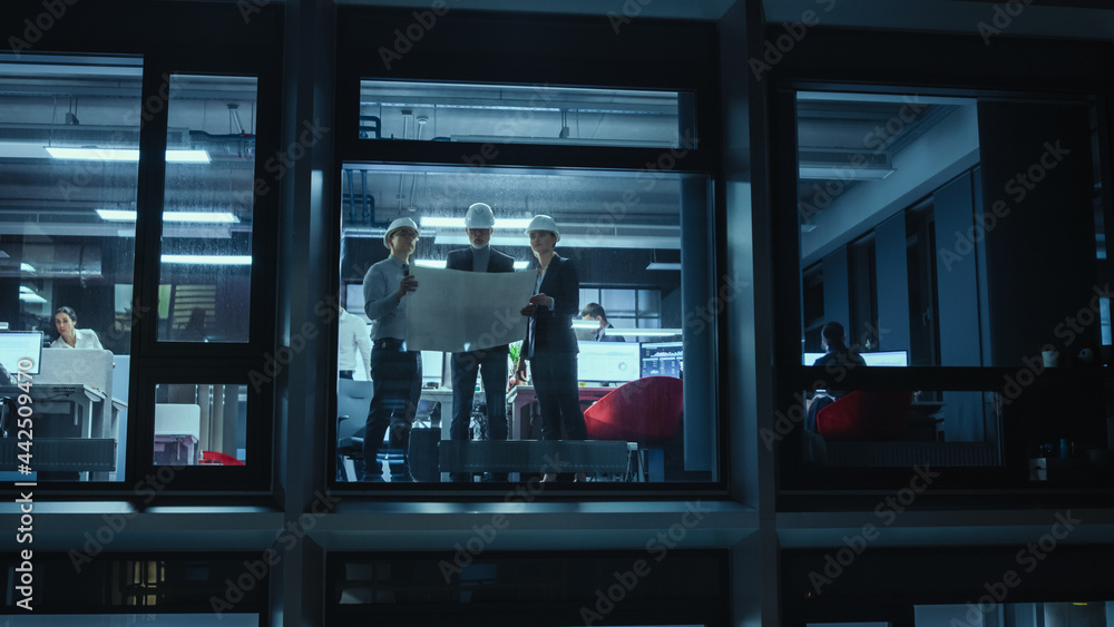 Poster Shot from Outside the Window: Architects and Engineers in Hard Hats Stand Next to a Window in Office Workplace and Look at Blueprints on Paper. Senior Specialists Explains the Job to His Apprentice