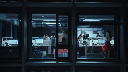 Shot from Outside the Window: Businessmen and Businesswomen Working in the Office. Managers and Specialists Doing Financial Business in the Evening. Employees Work on Computers and Delegate Tasks.