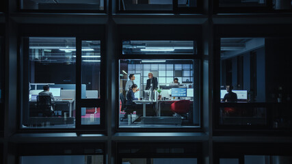 Shot from Outside the Window: Businessmen and Businesswomen Working in the Office. Managers Doing Financial Business in the Evening. Employees Work on Computers, Laptops, Mobile Phones, Tablets.