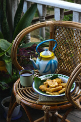 Morning tea with banana fritters, served on enamel tea set. Vintage photography with green plants background. Moody concept.