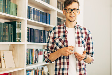 student in glasses and a plaid shirt drinks coffee and relaxes sitting at the table against the background of the wall with books
