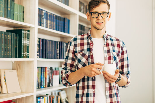 student in glasses and a plaid shirt drinks coffee and relaxes sitting at the table against the background of the wall with books