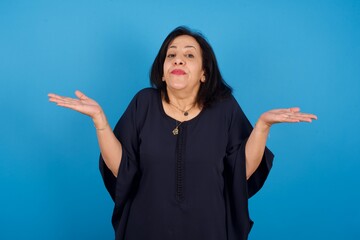 Maybe yes or no. Doubtful middle aged Arab woman standing against blue background shrugs shoulders in bewilderment, tries to make decision puzzled what he wants. Hesitation and uncertainty.