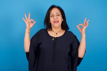 Glad middle aged Arab woman standing against blue background shows ok sign with both hands as expresses approval, has cheerful expression, being optimistic.