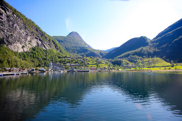Naklejka na ściany i meble Beautiful view of Geiranger town from the water on a summer day. Reflections of the mountains in the fjord. lush green landscape with peaks towering over the village. Norway.