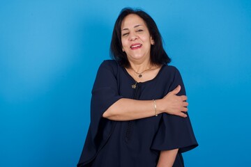 People, lifestyle, youth and happiness concept. Shy pretty middle aged Arab woman standing against blue background, feeling happy hugging herself.