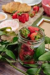 side view of fresh ripe strawberries in a glass jar and green leaves croissant on rustic wooden background