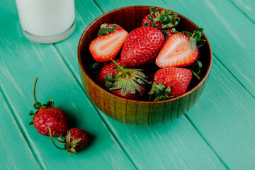side view of fresh ripe strawberries in a wooden bowl with a glass of milk on green wooden background