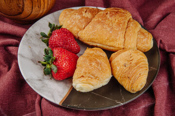 side view of fresh ripe strawberries with croissants on a plate on dark red fabric background