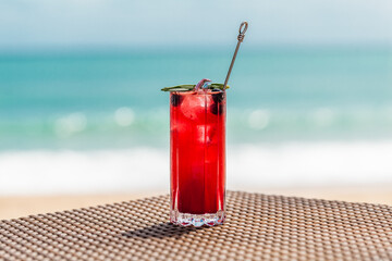 Fresh Red Berries Cocktail with Ice Cubes on Beach Table, Defocused Turquoise Sea on Background. Healthy Tropical Mocktail in Restaurant by the Sea. Ice Cold Drink in Paradise. Vacation Concept.