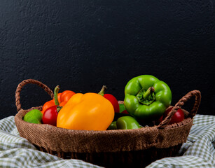 Fototapeta na wymiar side view of fresh vegetables colorful bell peppers tomatoes and cucumbers in a wicker basket on plaid fabric on black background