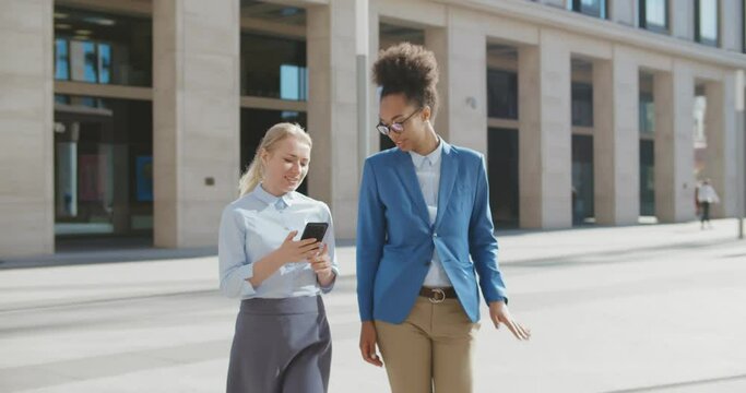Tall and short diverse women in office clothes walking outdoors and using phone.